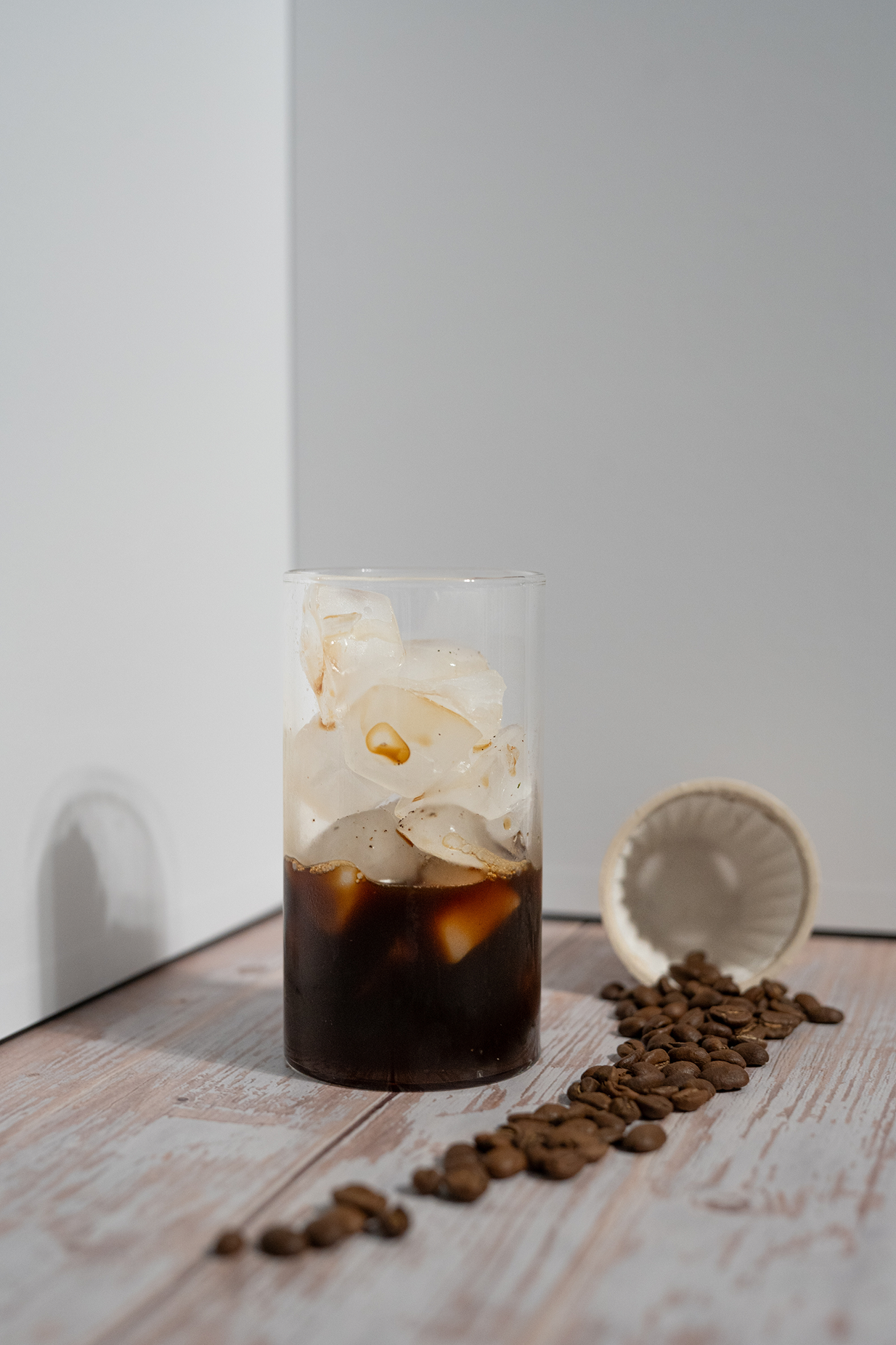Whole coffee beans beside an empty xPod filter container and a glass of iced black coffee